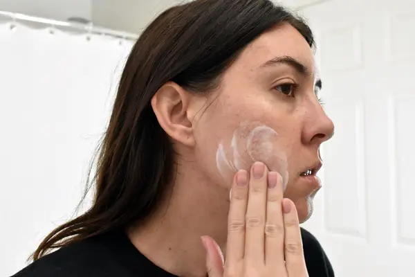 Person rubbing Differin Daily Deep Cleanser onto cheek with white shower curtain in background