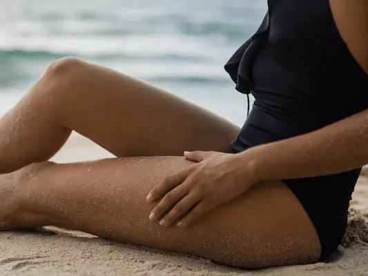 A tanned woman in a black bathing suit sitting on the beach with sand and her hand on her leg