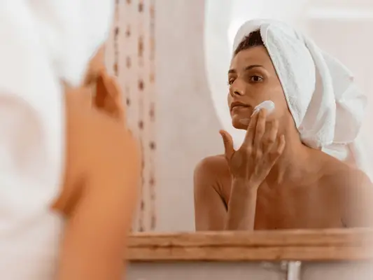 A woman looking in the mirror with her hair in a towel and applying a face mask