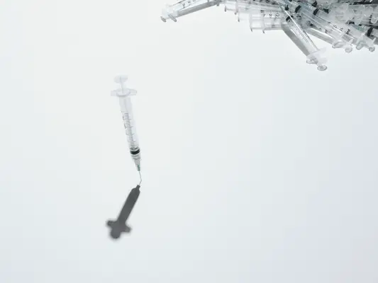 A syringe standing up on its needle in the center with a pile of syringes in the corner