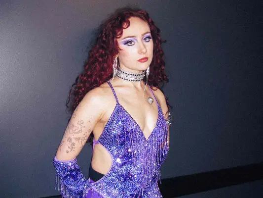 Chappell Roan wears a purple glitter fringe bodysuit and gloves with purple eyeshadow