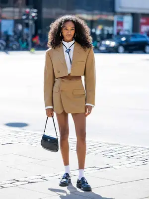 Woman wearing cropped blazer, matching skirt, white blouse, Prada bag, tie, and loafers, white socks, and hoop earrings