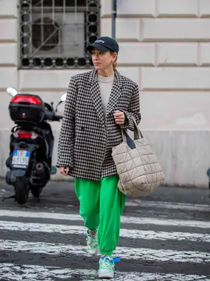 Woman wearing oversized houndstooth blazer, bright green baggy sweatpants. sneakers, baseball cap, crew neck sweater, and puffer bag