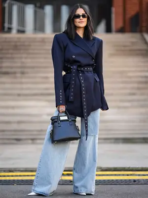 Woman wearing belted navy blazer, wide leg jeans, black handbag, pointed-toe white heels, and sunglasses