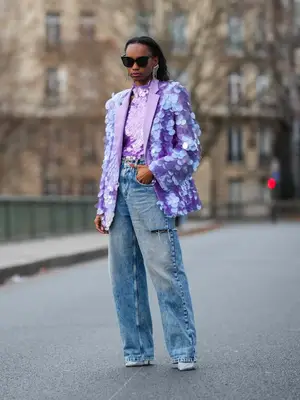 Woman wearing purple sequined blazer and top, wide leg jeans, sunglasses, pumps, and statement earrings