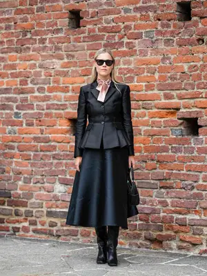 Woman wearing peplum blazer, black midi skirt, pink scarf, black knee-high boots, handbag, and sunglasses
