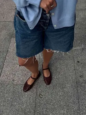 Close-up of person wearing brown ballet flats, long denim shorts, and blue cardigan
