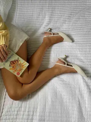 Close-up of person wearing cream-colored slingback heels, yellow sweater, and beige linen shorts while holding book