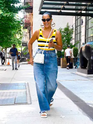 Tracee Ellis Ross wears striped tank top, wide leg jeans, white Birkenstock sandals, white crossbody handbag, angular sunglasses, and oversized hoop earrings