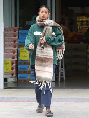 Willow Smith wears a green Billabong sweatshirt, an oversized striped scarf, jeans, and brown Birkenstocks with socks