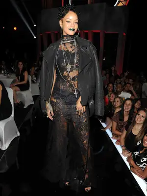 Rihanna wears a black lace dress, leather jacket, layered necklaces, hoop earrings, and bantu knots