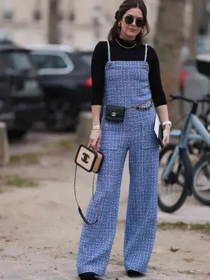 Paola Alberdi wears a blue tweed jumpsuit, black turtleneck, black belt bag, carries a black and beige Chanel bag, black shades, and heels 