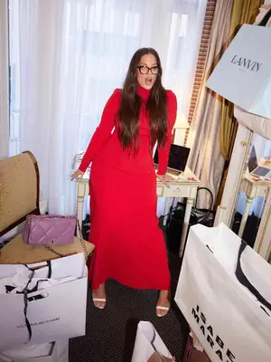 Ashley Graham wears a red turtleneck dress, glasses, and white heels with purple quilted Chanel bag on chair