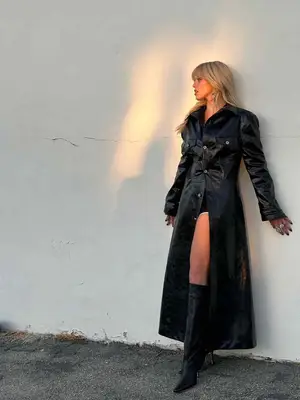 Renee Rapp standing against a white wall wearing a black trench coat and tall boots