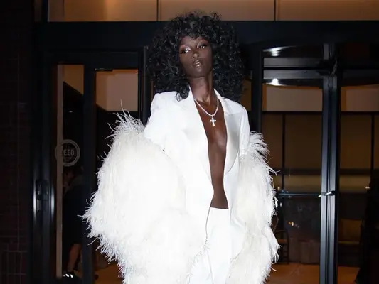 Anok Yai wears a white suit with open blazer, silver cross necklace, and feathered coat