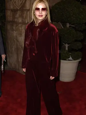 Priscilla Presley wears a red velvet suit, fur scarf, and square sunglasses