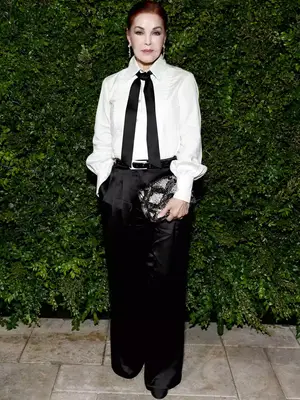 Priscilla Presley wears a puff-sleeved white button down, black trousers, a Chanel bag, and a black tie