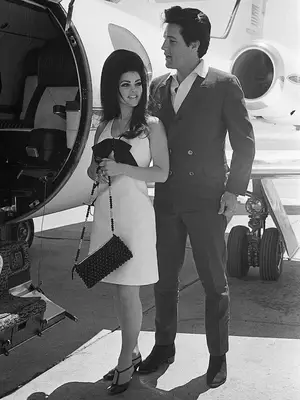 Priscilla Presley wears a white shift dress with bow, studded shoulder bag, and T-strap heels while boarding a plane with Elvis