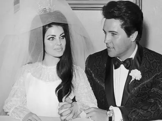 Priscilla Presley wears a puff-sleeved white gown, wedding veil, and crown while sitting with husband Elvis
