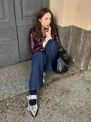 Laufey wears a burgundy structured jacket, wide leg jeans, silver buckled flats, and black slouchy handbag with chain strap