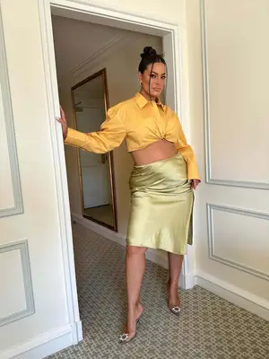 Ashley Graham wears a yellow button-down shirt, sage green satin slip skirt, clear pumps, and updo hairstyle