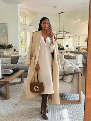 Jasmine Tookes wears a white knit dress with plunge neckline, cream-colored coat, brown purse with silver logo, and croc boots