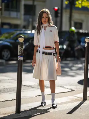 Jenny Suet In Tsang wears a white polo with blue and red stripes, white pleated midi skirt, exposed briefs, loafers and socks, and a peach handbag with a white necktie as scarf