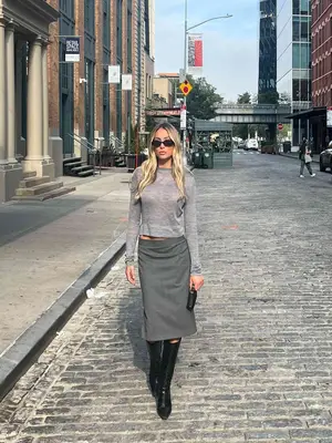 Alex Earle wears a long-sleeve gray shirt, gray midi skirt, sunglasses, and black knee-high boots 