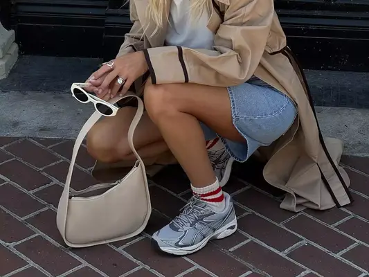 Close-up of woman wearing trench coat, white shirt, denim shorts, sneakers with striped socks, beige handbag, and white sunglasses