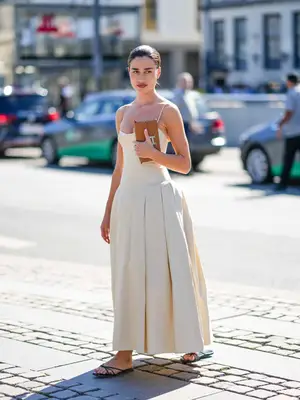 Woman in a cream-colored pleated drop-waist, spaghetti strap maxi dress, brown clutch, and black flat sandals