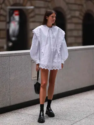 Woman in a white long sleeve shirt dress with an oversized lapel and eyelet details, black platform loafers and socks, and black handbag