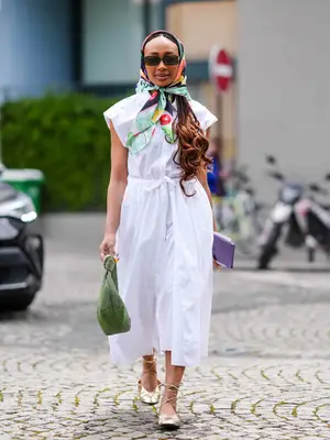 Woman wearing a white cap-sleeve midi shirt dress, headscarf, sunglasses, green handbag, and gold ballet flats with ties