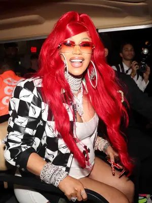 Cardi B smiling at camera with red hair, checkered leather jacket, large hoops, and red y2k inspired sunglasses