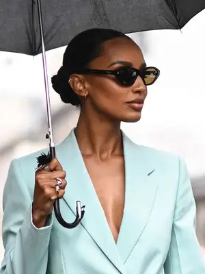 Jasmine Tookes walking outside holding umbrella with blue blazer, hair in bun, and oval sunglasses