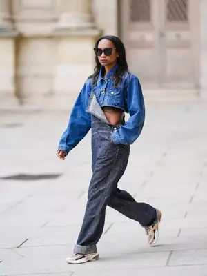 Model walking and wearing overalls, a denim jacket, sunglasses, and sneakers