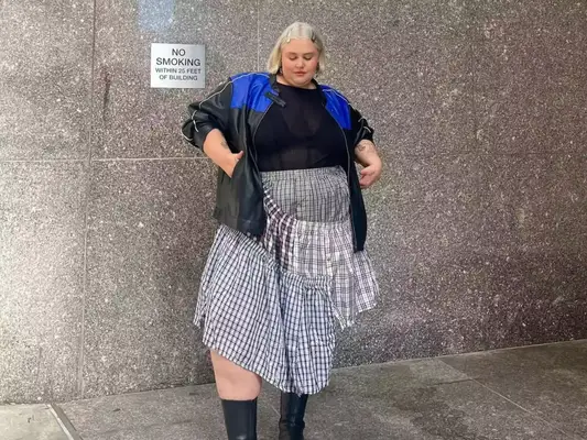 A content creator poses for a photo while wearing a plaid skirt, sheer black top and a moto jacket