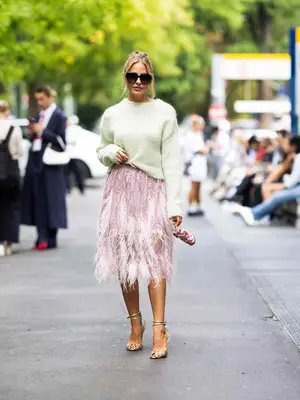 Ginta wears a mint green sweater, pink feathered skirt, gold pumps, and oversized sunglasses
