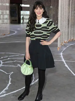 Zooey Deschanel wears a green and black short-sleeved sweater, black voluminous skirt, and green handbag with black tights and boots