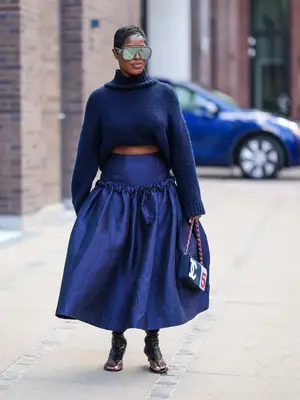 Woman wears navy mock neck sweater, voluminous blue skirt, Chanel logo bag, shield sunglasses, and open-toe black heels