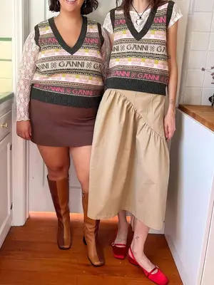 Two women wearing pink and green striped Ganni logo sweaters, white lacy shirts, and neutral colored skirts