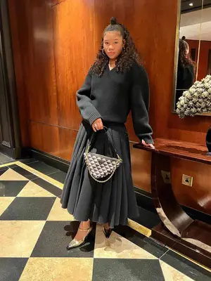 Storm Reid wears a dark V-neck sweater, pleated maxi skirt, Prada logo bag, and Mary Jane heels