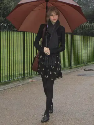 Taylor Swift wearing black tights, black flats, a black mini dress, scarf, and brown leather bag while walking with a brown umbrella in London