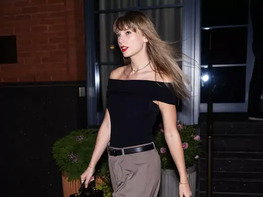 Taylor Swift wears a black boat neck top, beige pants, and minimal jewelry