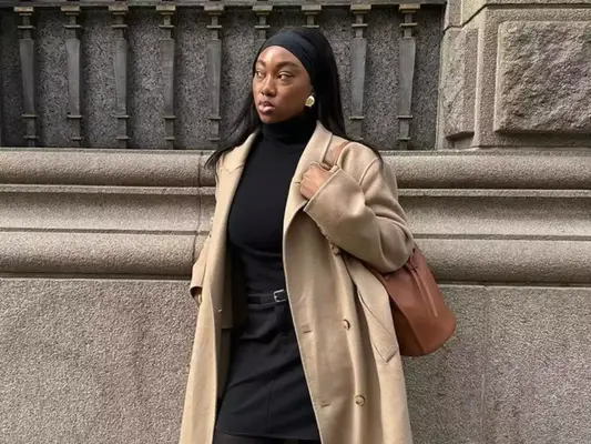 Woman posing while wearing a light brown trenchcoat, black turtleneck, black skirt, a belt, and a soft headband. 