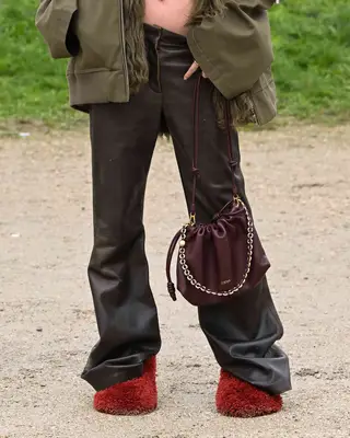 Close-up of person wearing shearling red shoes, oxblood handbag, black leather pants, and taupe jacket