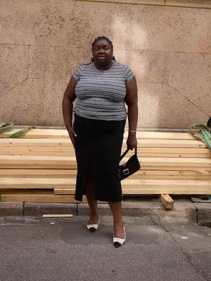 Abisola Omole wears a striped t-shirt, mid-length black skirt, and contrast-toe shoes.