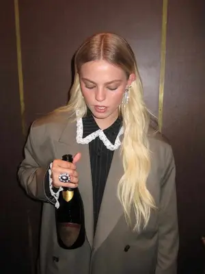 Renee Rapp wears a collared shirt and blazer with waterfall earrings. She holds a bottle of Champagne
