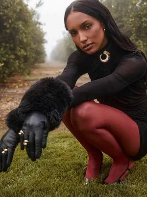 Jasmine Tookes wears a gold brooch, black ruched dress, red tights, black slingback heels, and fur-trim leather gloves with rings overtop