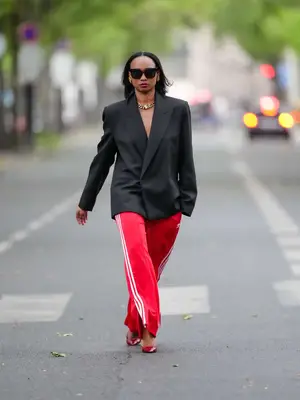  Emilie Joseph wears sunglasses, earrings, red lipstick, a black oversized / structured virgin wool blend Blazer with lapel-collar by Victoria Beckham for Mango, Adidas Classics oversized tracksuit bottoms / track pants in red with stripes, a golden chunky necklace / chain by Merbabe Greece, shoes : Newton satin pointy black pumps with high heels by Saint Laurent,