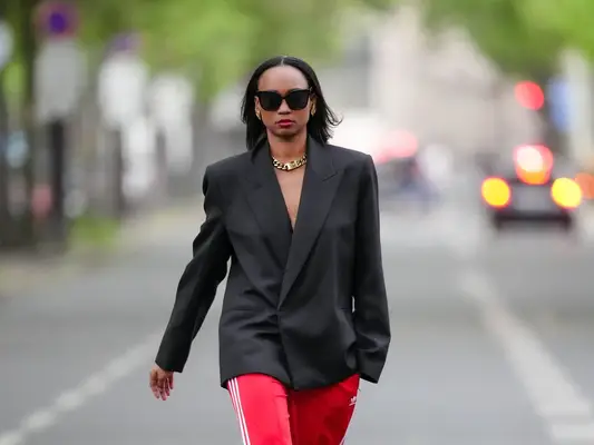 Emilie Joseph wears sunglasses, earrings, red lipstick, a black oversized / structured virgin wool blend Blazer with lapel-collar by Victoria Beckham for Mango, Adidas Classics oversized tracksuit bottoms / track pants in red with stripes, a golden chunky necklace / chain by Merbabe Greece, shoes : Newton satin pointy black pumps with high heels by Saint Laurent,